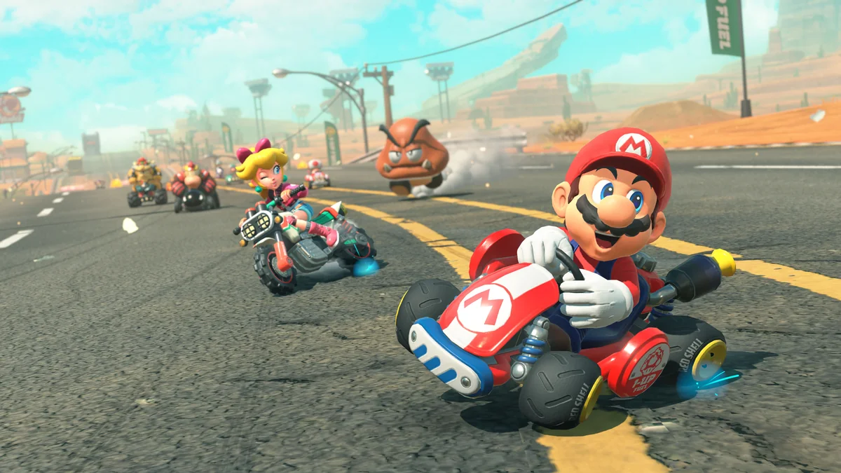 Mario derrapando en un kart rojo por un circuito desértico, con Peach en moto y un Goomba al fondo