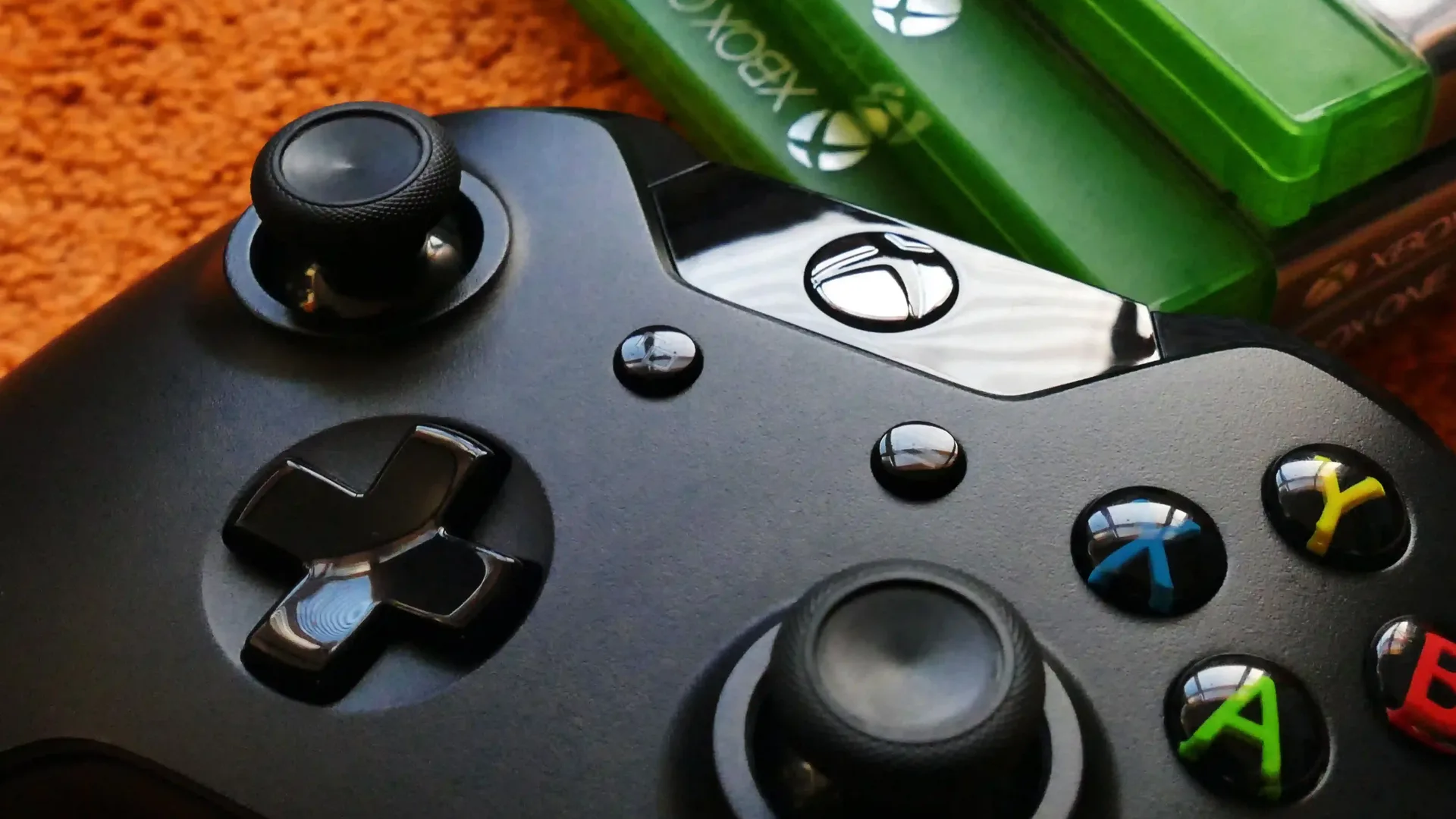 Mando negro de Xbox sobre una alfombra naranja, con cajas de juegos al fondo